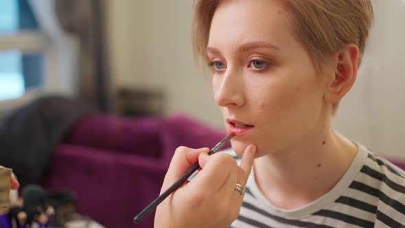 a Young Woman with a Short Haircut Sits and a Makeup Artist Applies Lipstick to Her Lips with a