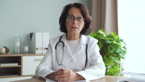 Portrait of Mature Female Doctor Is Talking Sitting at Table in Clinic Room.