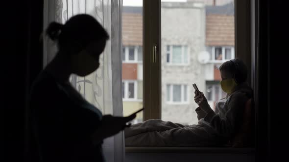 Woman and Teenager Holding Smartphone. Both in Protective Masks Opposite the Window. Quarantine 2020