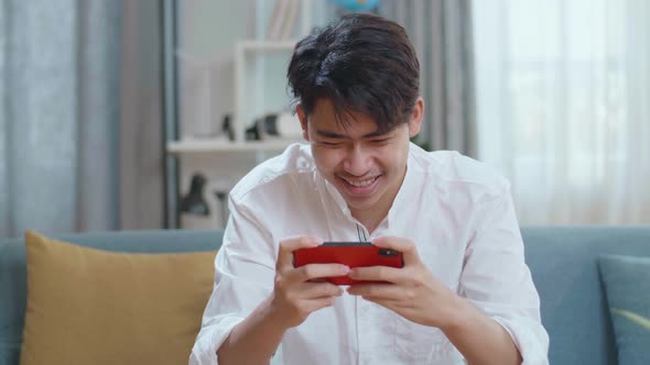 Asian Man Playing Game On Smartphone And Smiling While Sitting On Sofa In The Living Room