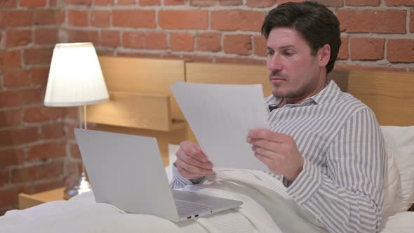 Middle Aged Man with Laptop Reading Documents in Bed