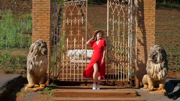 Glamorous Stylish Woman in a Red Dress Posing Near Wrought Iron Gates with Lion Sculptures in a