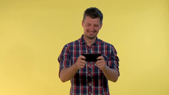 Positive Young Man Enjoy Playing Online Game on Smartphone on Yellow Background