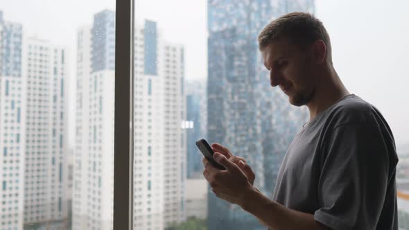 Caucasian Man Browsing His Smartphone While Enjoying Urban View From His Skyscraper Apartment