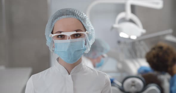 Portrait of Young Confident Female Dentist in Protective Cap Gloves and Mask Posing in Dental Office