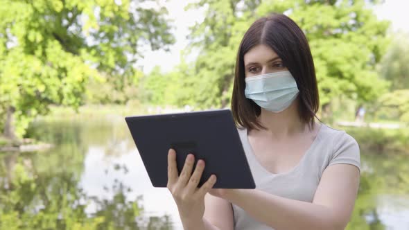 A Young Caucasian Woman in a Face Mask Works on a Tablet in a Park  a Pond and Trees in Background