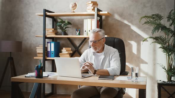 Senior Male Saying Hello to Business Partner Using Online Video Call on Notebook While Sitting at