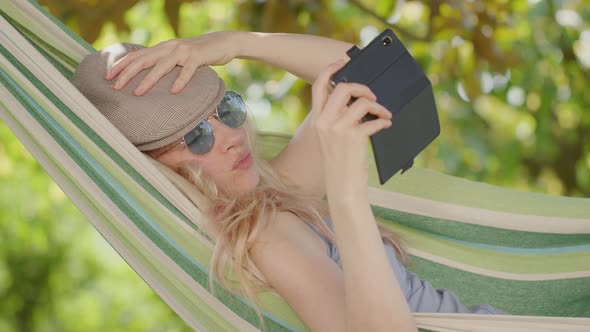 Smiling blonde woman with sunglasses using smartphone, lying relaxing on the hammock in the garden