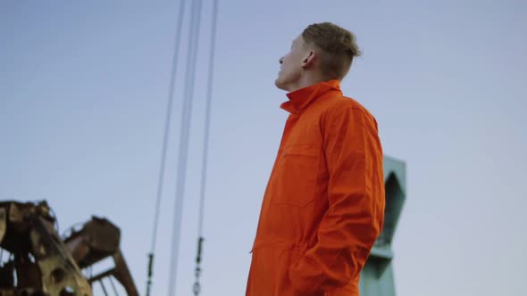 Handsome Young Container Warehouse Worker in Orange Uniform Standing By the Ship at the Harbor and