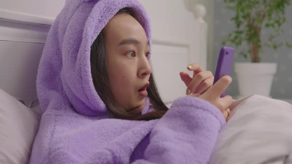 Young Adult Asian Woman Reading Good News on Cell Phone Lying in Bed