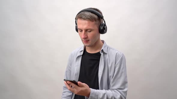 A Young Man on a Gray Background in Headphones is Typing Text on a Smartphone