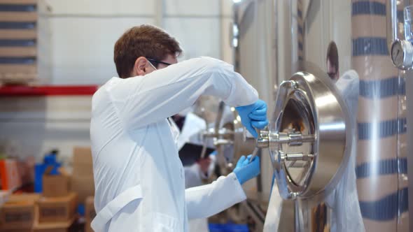 Concentrated Experienced Brewing Technician in Coat and Mask Turning Handle of Brewing Equipment