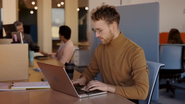 Young Businessman Sitting at Desk and Typing on Laptop Computer in Contemporary Corporation Office