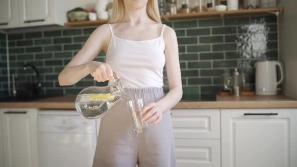 Young Beautiful Woman Pours Water with Lemon Into a Glass and Drinks