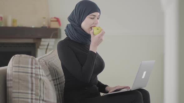Young Muslim Woman Working or Chatting By Laptop and Biting Juicy Apple