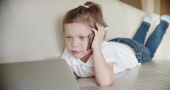 A Little Girl Lies at Home on the Couch and Looks Into a Laptop