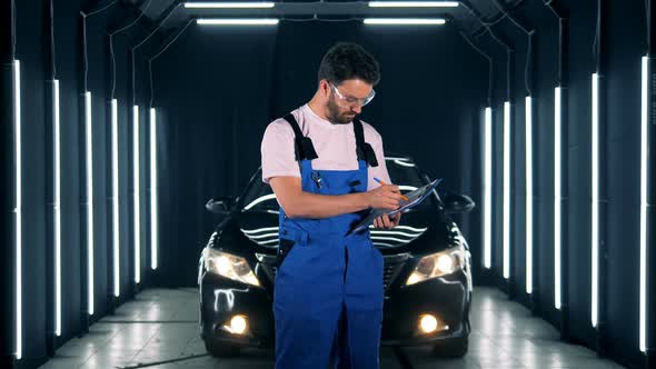 Repairman Writes on a Clipboard on a Car Background. Worker Stands in a Garage, Holding a Clipboard