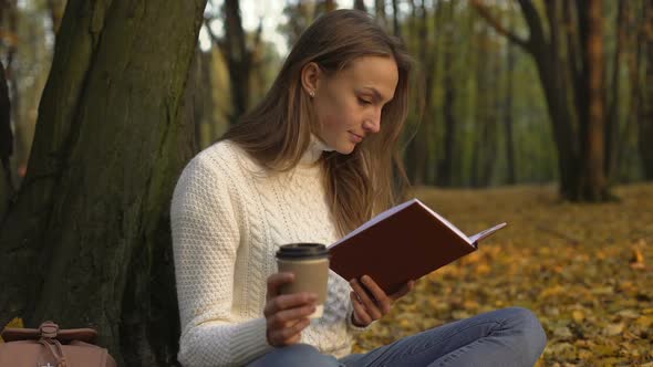 Female Student Reading Book Sitting in Park on Warm Autumn Day