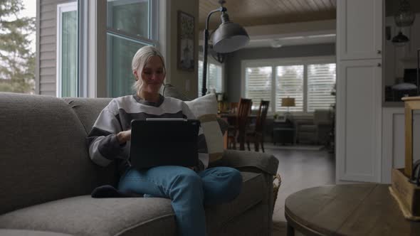 Woman sitting on the couch in front of a bay window, drinking coffee while scrolling on a tablet wit