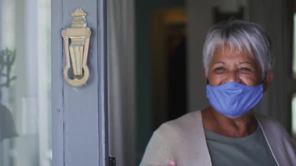 Senior mixed race woman wearing face mask opening front door waving greeting