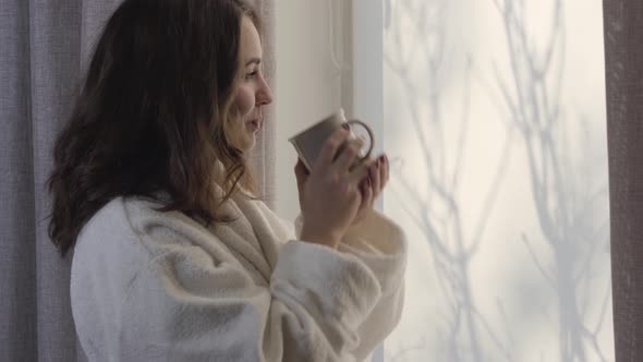 Close-up Side View of Young Beautiful Woman Drinking Coffee at the Window and Fixing White Bathrobe