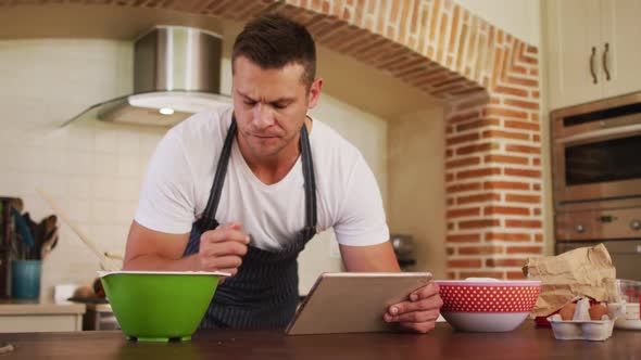 Caucasian man wearing apron smiling while using digital tablet in the kitchen at home