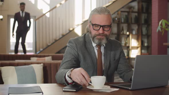 Senior Businessman Drinking Coffee and Working on Laptop in Restaurant