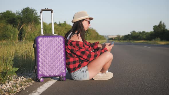 Traveler Woman with Suitcase Sits on Road and Waiting for a Car To Travel, Pretty Young Woman