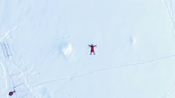 Aerial Camera Rises Up, a Top View of a Girl in a Red Fluff Lying and Making a Snow Angel