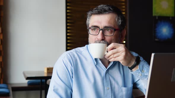 Senior Man Drinks Coffee Sitting in a Cafe with a Laptop