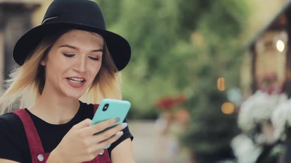 Portrait of Smiling Young Pretty Blond Girl Wearing a Black Hat Using Her Smartphone Standing at the