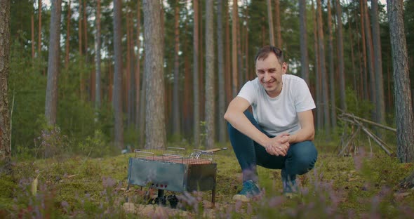 Cheerful Adult Man Is Sitting Near Chargrill with Meat and Sausages in Forest, Smiling and Enjoying