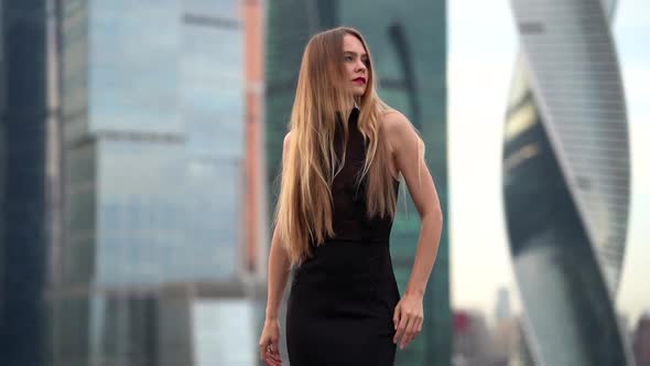 a Woman with Long Blonde Hair and a Black Evening Dress Stands Against a Background of High-rise