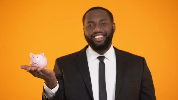 Thrifty Afro-American Man in Suit Putting Coin in Piggy Bank, Savings, Deposit