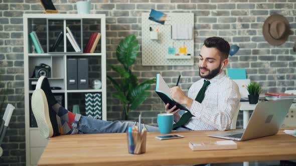 Cheerful Guy Listening To Music Through Earphones Writing in Notebook at Work