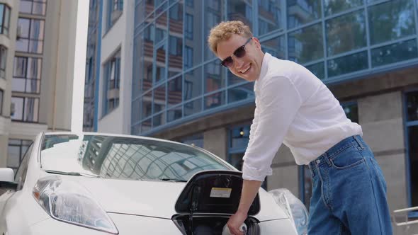 Stylish Modern Young Curly Man Stands By the Battery and Holds a Connected Charger