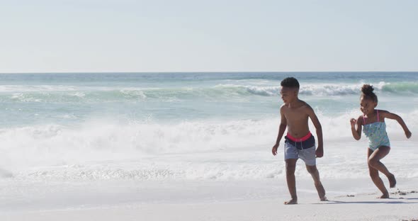 Smiling african american siblings running and wearing swimming suits on sunny beach