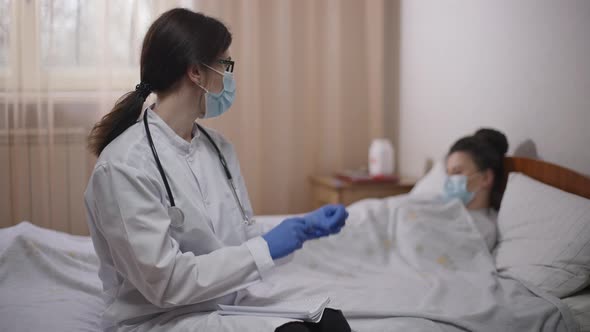 Concentrated Female Doctor Putting on Gloves Looking Back at Blurred Ill Woman Lying in Bed in