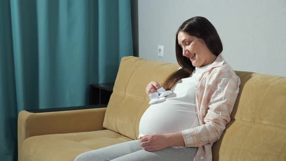 Pregnant Woman Holding White Booties in Hand Stroking Belly While Sitting on the Couch