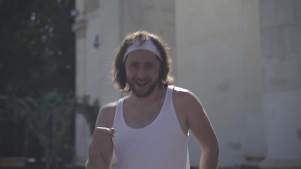 Handsome Positive Young Sportsman Walking Fast in Sunlight Outdoors. Smiling and Winking at Camera