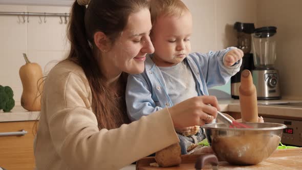 Smiling Young Mother with Little Baby Son Making Bread Dough in Bowl on Kitchen