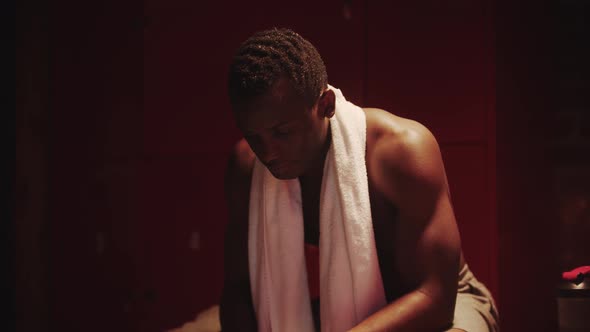 Darkskinned Young Man Sits in a Locker Room with a Towel Around His Neck