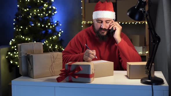 Small Business Owner Bearded Indian in Santa Claus Costume in Home Office Takes an Order for Goods