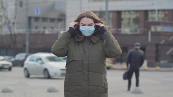 Middle Shot of Young Brunette Woman Standing on City Street and Putting on Protective Mask