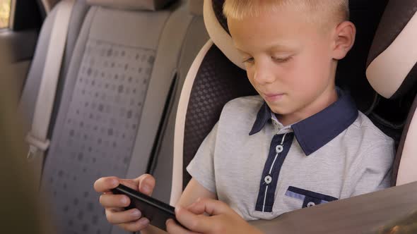 A Cute Little Boy is Sitting in a Car in a Car Seat Playing with His Phone