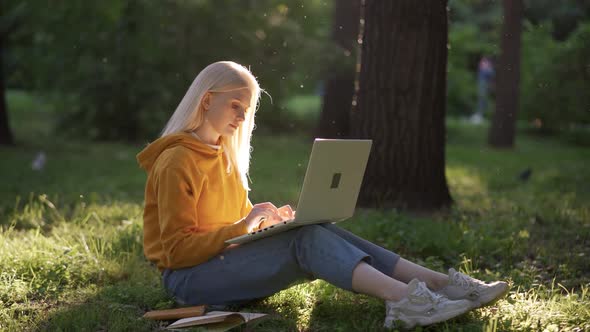 A Young Woman in Jeans and a Bright Yellow Sweatshirt with a Laptop Sits on the Green Grass
