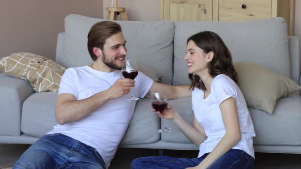 Happy couple sitting, relaxing on floor in living room, drinking red wine.