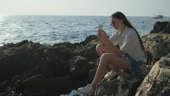 Woman is Taking Photo of Seascape Sitting on Stones