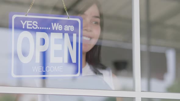 Asian woman business owner hanging blue open sign label on entrance door of coffee shop cafe