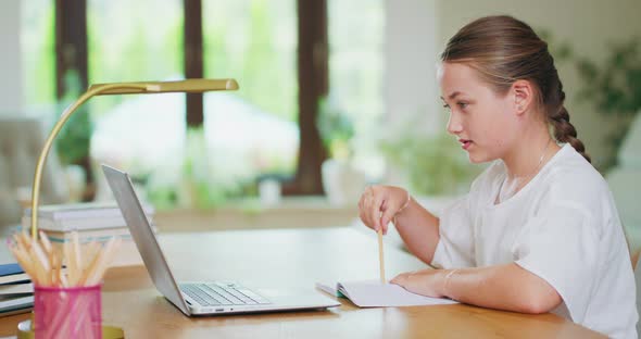 Closed Focused Teen Girl at the Desk Writes Tasks in Notebook and Checks Them on Laptop Books alt
